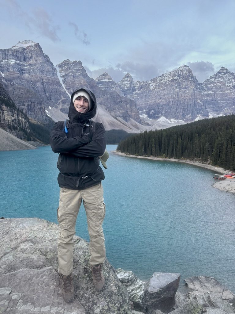 Anthony on a rock above a turquoise mountain lake, peaks behind