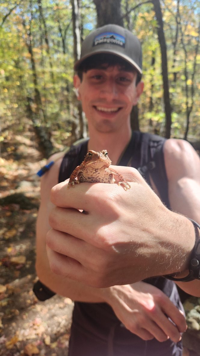 Anthony on a wooded trail, smiling behind a small toad on his hand